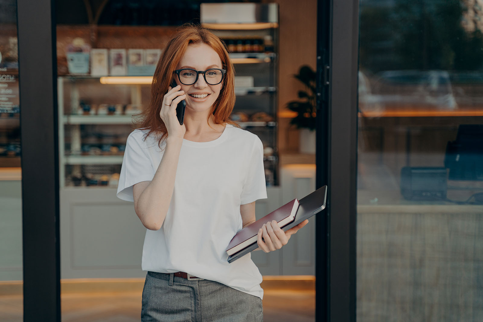 Smiling woman with laptop and notebook in hand enjoying talk on cellphone, consulting client or customer while standing near coffee shop, happy young redhead businesswoman talking smartphone outdoor